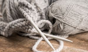 Zwei weiße Stricknadeln liegen in einem halbgestrickten Stück aus weichem, hellgrauem Garn, daneben liegt auf einer Holzoberfläche ein Garnknäuel.