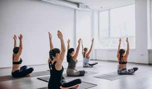 Fünf Personen sitzen in einem hellen Studio auf Yogamatten, blicken nach vorne, strecken die Arme über den Kopf und üben eine Yoga-Pose. Der Raum hat große Fenster und weiße Wände, was eine ruhige und luftige Atmosphäre schafft.
