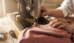 Nahaufnahme von Händen, die rosa Stoff durch eine Vintage-Nähmaschine führen. Garnrollen und Nähwerkzeuge liegen daneben auf dem Tisch. Warmes, sanftes Licht unterstreicht den Herstellungsprozess.