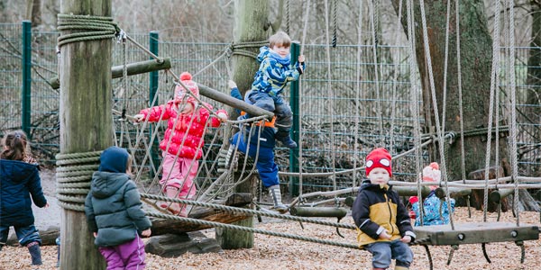 Kinder in Winterkleidung spielen auf einem hölzernen Klettergerüst mit Seilen auf einem Spielplatz einer Kindertagesstätte, umgeben von Bäumen und einem Drahtzaun. Der Boden ist mit Holzspänen bedeckt und schafft so eine sichere und spielerische Kinderbetreuungsumgebung.