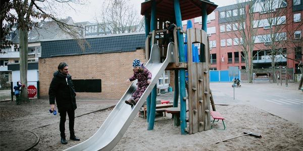 Ein Kind in einem gemusterten Schneeanzug rutscht auf einer Spielplatzrutsche einer der örtlichen Kindertageseinrichtungen hinunter, während ein Erwachsener in der Nähe steht und zusieht. Der Sandspielplatz ist im Hintergrund von Gebäuden und Bäumen umgeben.