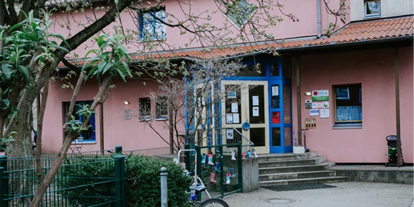 Ein rosafarbenes Gebäude mit blauen Türen und Fenstern, einem Ziegeldach und Schildern in der Nähe des Eingangs lässt vermuten, dass es sich um eine der örtlichen Kindertageseinrichtungen handeln könnte. Ein Baum, ein Fahrrad, ein grüner Zaun und Stufen führen zur Tür hinauf.