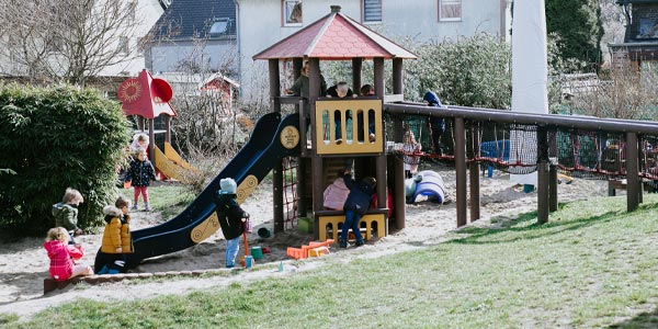 Kinder spielen an einem sonnigen Tag auf einem Spielplatz der Kindertageseinrichtungen und vergnügen sich mit Rutsche, Klettergerüst und Sandkasten. Einige Kinder klettern, andere spielen im Sand, während einige die Rutsche benutzen. Im Hintergrund sind Häuser und Bäume zu sehen.