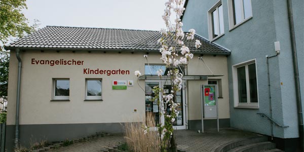 Eingang eines Gebäudes mit einem Schild mit der Aufschrift „Evangelischer Kindergarten“ an der Wand, einem blühenden Baum davor, einem gepflasterten Weg, der zu Glastüren führt, und Fenstern an beiden sichtbaren Wänden, die einen einladenden Ort für die Kinderbetreuung widerspiegeln.