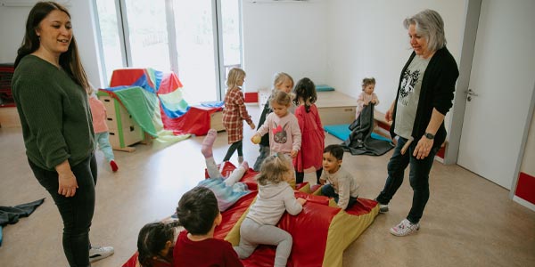Mehrere kleine Kinder spielen auf bunten Matten in einem hellen Klassenzimmer der Kindertageseinrichtung, während zwei Frauen sie beaufsichtigen. Der Raum verfügt über große Fenster und überall stehen verschiedene Spielgeräte herum.
