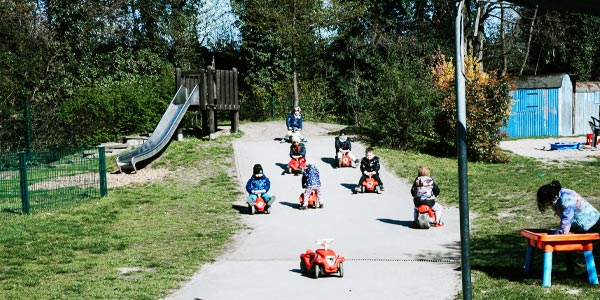 Mehrere kleine Kinder aus einem Kindergarten fahren mit kleinen roten Spielzeugautos auf einem gepflasterten Weg in einem Park. Im Hintergrund sind eine Rutsche und grüne Bäume zu sehen. Ein Kind spielt an einem Tisch auf der rechten Seite dieser Kinderbetreuungseinrichtung.