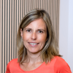 Eine Frau mit schulterlangem blondem Haar im korallenfarbenen Oberteil lächelt vor hellem Hintergrund.