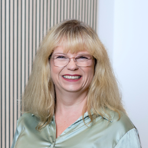 Eine Frau mit langen blonden Haaren und Brille lächelt in die Kamera. Sie trägt eine hellgrüne Satinbluse.