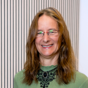Eine Frau mit langen braunen Haaren, Brille, grünem Shirt und schwarzer Kette lächelt.