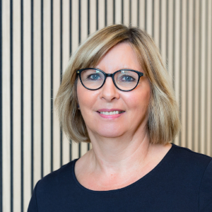 Eine Frau mit kurzen blonden Haaren und Brille lächelt in die Kamera. Sie trägt ein dunkles Oberteil.