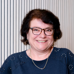 Eine Frau mit kurzen, dunklen Haaren und Brille lächelt in die Kamera. Sie trägt ein marineblaues Oberteil mit Ösendetails und eine goldene Halskette.