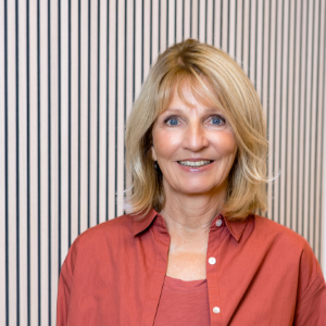 Eine lächelnde Frau mit blonden Haaren und einem korallenfarbenen Hemd steht vor einer weißen Wand mit vertikalen schwarzen Streifen.