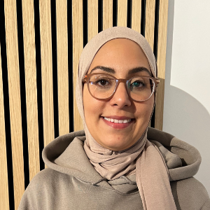 Eine Frau mit Brille und beigem Hijab lächelt in die Kamera. Sie trägt einen Kapuzenpullover und steht vor einer hellen Holzlamellenwand.