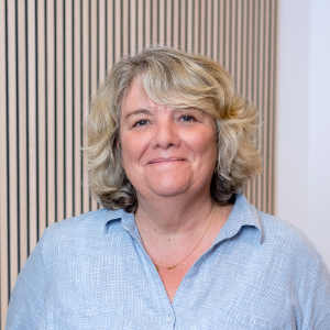 Eine Frau mit welligem blondem Haar, einem hellblauen Hemd mit Knöpfen und einer goldenen Halskette lächelt in die Kamera.