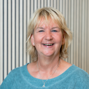 Eine lächelnde Frau mit kurzen blonden Haaren, einem türkisfarbenen Oberteil und einer silbernen Halskette vor einem hellen Hintergrund.