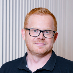 Ein Mann mit kurzen roten Haaren, heller Haut und Brille, der ein schwarzes Poloshirt trägt, lächelt leicht.