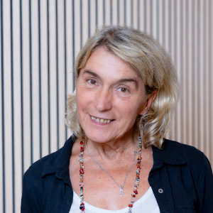 Eine lächelnde Frau mit welligem blondem Haar, Teil der Mitarbeitendenvertretung, trägt ein dunkles Hemd über einem weißen Oberteil und eine bunte Perlenkette.