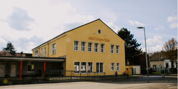 Ein gelbes, zweistöckiges Schulgebäude mit großen Fenstern und dem Namen Astrid-Lindgren-Schule an der Fassade, mit Programmen des Offenen Ganztags, an einem bewölkten Tag mit ein paar Menschen in der Nähe des Eingangs und der Straße.