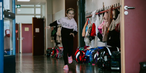 Ein lächelndes Mädchen läuft in einem Flur an einer Reihe bunter Rucksäcke und Mäntel vorbei, die an Haken hängen und den lebendigen Geist einer Offenen Ganztags-Schulumgebung einfangen.