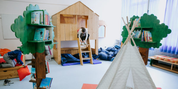 Ein gemütlicher Offener-Ganztag-Klassenraum mit hölzernen Spielgeräten, darunter ein kleines Haus und ein Tipi-Zelt. Baumförmige Bücherregale beherbergen bunte Bücher, und ein Kind klettert über eine Leiter in das Holzhaus.