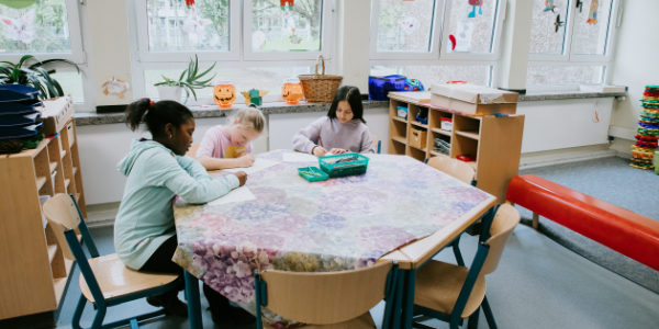 Drei Kinder sitzen in einem Klassenzimmer des Offenen Ganztags um einen Tisch und zeichnen oder schreiben auf Papier. Der Raum ist mit Regalen voller Materialien, dekorierten Fenstern und einer entspannten, hellen Atmosphäre ausgestattet.