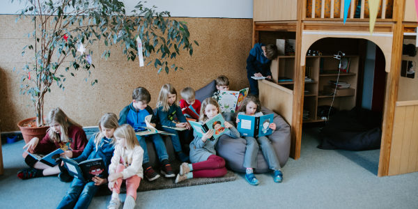 Eine Gruppe von Kindern sitzt auf dem Boden und Sitzsäcken und liest gemeinsam Bücher in einem gemütlichen Offenen Ganztag-Klassenzimmer mit einer Spielhausstruktur und einem Topfbaum in der Nähe.