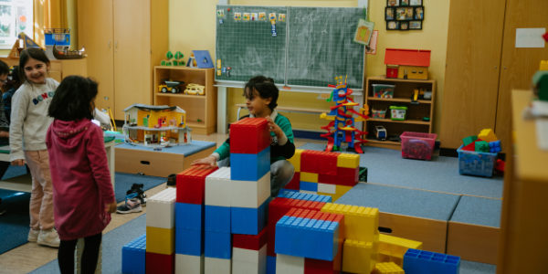 Drei kleine Kinder spielen in einem Klassenzimmer des Offenen Ganztags mit großen bunten Bauklötzen. Regale mit Spielzeug, eine Tafel und lehrreiche Dekorationen schaffen eine lebendige und einladende Lernumgebung.