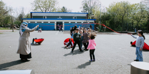 Kinder spielen am Offenen Ganztag auf einem Schulhof Seilspringen, beaufsichtigt von einem Erwachsenen mit Kopftuch. Andere Kinder haben in der Nähe Spaß mit roten Kreiselspielzeugen. Im Hintergrund sind das Schulgebäude und Bäume zu sehen.