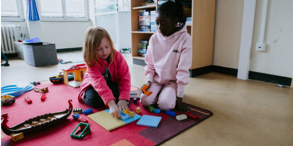 Beim Offenen Ganztag sitzen zwei Kinder auf einem roten Teppich und bauen mit bunten Plastikklötzen. Umgeben von Spielzeug und Spielen sind beide in diesem einladenden Raum ganz auf ihr kreatives Spiel konzentriert.