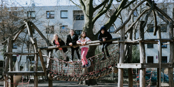 Vier Kinder vom Offenen Ganztag stehen und sitzen auf einer Holz- und Seilbrücke auf einem Spielplatz und lächeln in die Kamera. Im Hintergrund sind Bäume und Wohnhäuser zu sehen.