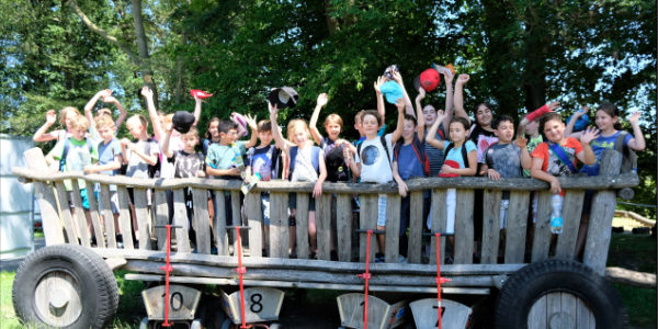 Eine Gruppe von Kindern des Offenen Ganztags steht gemeinsam auf einer großen Holzkonstruktion im Freien und winkt und lächelt in die Kamera. Die Umgebung ist sonnig, im Hintergrund sind Bäume und Grün zu sehen.