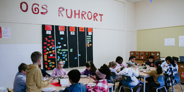 Eine Gruppe von Kindern sitzt an Tischen, isst und unterhält sich in einem Klassenzimmer, das mit bunten Notizen und dem an die Wand gemalten Schriftzug „OGS Ruhrort“ dekoriert ist und die lebendige Atmosphäre eines Programms des Offenen Ganztags widerspiegelt.