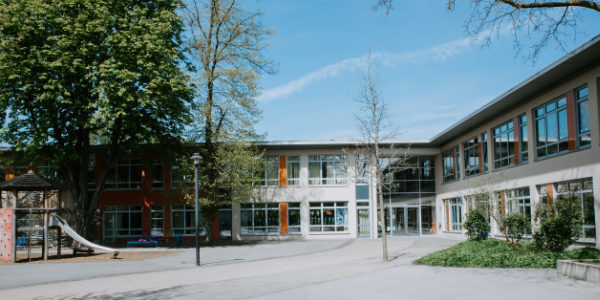 Ein modernes Schulgebäude mit großen Fenstern und zwei Stockwerken sowie Möglichkeiten für den Offenen Ganztag ist von Bäumen und einem Außenspielplatz unter einem klaren blauen Himmel umgeben.