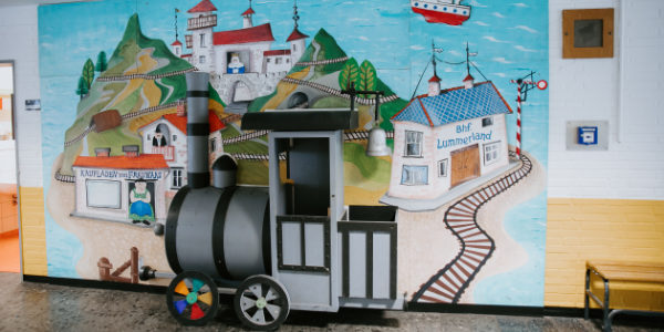 Ein kleines dekoratives Eisenbahnmodell steht vor einem farbenfrohen Wandgemälde mit Comic-Gebäuden, Gleisen und Hügeln und schafft so eine verspielte und skurrile Szene im Innenbereich – perfekt, um jeden Raum für den Offenen Ganztag aufzuhellen.
