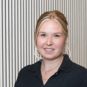 Eine Frau mit blondem Haar zu einem tiefen Dutt, einem schwarzen Top und zarten Goldketten steht vor vertikalen schwarz-weißen Streifen.