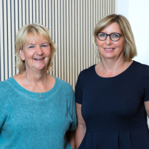Zwei Frauen mittleren Alters mit blonden Haaren stehen lächelnd nebeneinander. Als freundliche Ansprechpartnerin trägt die eine einen blaugrünen Pullover und die andere eine Brille und ein marineblaues Kleid und posiert vor einer hellen, vertikal gestreiften Wand.