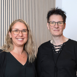 Zwei Frauen mit Brille stehen nebeneinander und lächeln in die Kamera. Mit ihren unterschiedlichen Stilen wirken sie nahbar – perfekte Ansprechpartnerinnen. Die gestreifte Wand im Hintergrund verleiht ihrem Porträt eine moderne Note.
