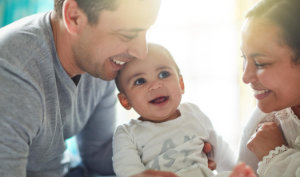 Ein lächelndes Baby sitzt zwischen zwei Erwachsenen, die sich eng aneinanderlehnen und das Kind herzlich anlächeln. Sonnenlicht erhellt sanft ihre Gesichter und schafft eine fröhliche und liebevolle Atmosphäre.