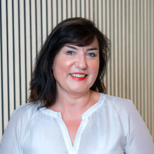 Eine Frau mit schulterlangem dunklem Haar und rotem Lippenstift, spezialisiert auf Kinderbetreuung, lächelt in die Kamera. Sie trägt eine weiße Bluse und steht vor einem Hintergrund aus vertikalen schwarz-weißen Streifen.