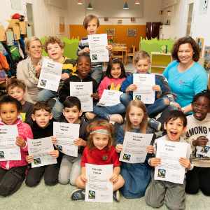 Eine Gruppe lächelnder Kinder, die drinnen sitzen und stehen, hält stolz ihre Zertifikate hoch. Zwei Erwachsene sitzen mit ihnen in dem farbenfrohen, einladenden Raum und feiern ihre „Zertifizierte Faire Kita“ bei der Duisburger Lutherkirche.