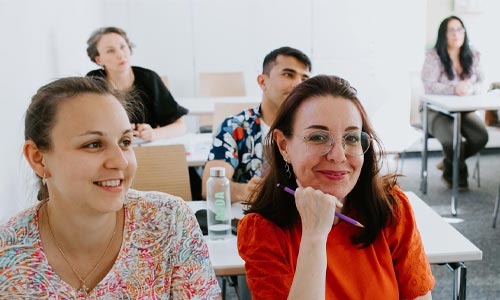 Eine Gruppe Erwachsener sitzt in einem hellen Klassenzimmer an ihren Tischen und hört aufmerksam zu. Zwei Frauen in der ersten Reihe lächeln, eine hält einen Bleistift in der Hand, während andere dahinter sitzen und sich auf den Unterricht konzentrieren.