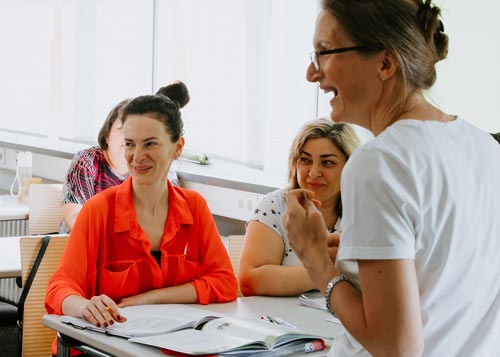 Drei Frauen sitzen mit offenen Büchern an einem Tisch, lächeln und unterhalten sich mit einer stehenden Frau in einem weißen Hemd, während sie in einem hellen Klassenzimmer ihre beruflichen Sprachkenntnisse verbessern.