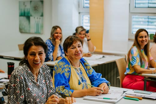 Eine Gruppe Frauen sitzt lächelnd an ihren Tischen in einem Elternintegrationskurs-Klassenzimmer und blickt in die Kamera. Sie haben Notizbücher und Stifte vor sich und wirken aufmerksam und glücklich, während helles Tageslicht durch die Fenster fällt.