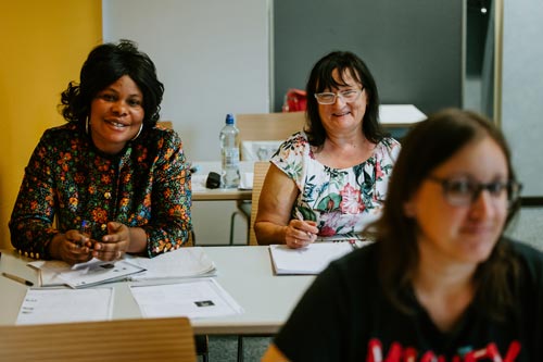 Drei Frauen sitzen während eines Integrationskurses an ihren Tischen in einem Klassenzimmer. Zwei lächeln in die Kamera, vor ihnen liegen Hefte und Papiere. Sie verkörpern damit den Geist von Integration und Deutschlernen. Eine Frau ist im Vordergrund leicht unscharf.