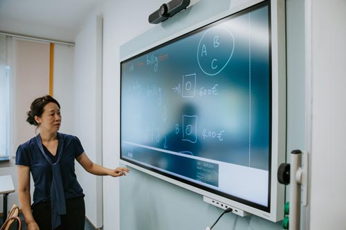 Eine Frau steht neben einem großen interaktiven Bildschirm, auf dem mathematische Diagramme, Formeln und Eurobeträge angezeigt werden, und zeigt auf den Bildschirm, als würde sie im Unterricht ein Integrationskursangebot erklären.