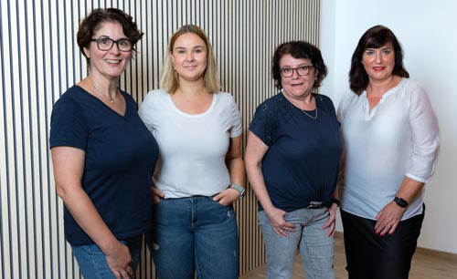 Vier Frauen stehen nebeneinander in einem Raum und lächeln in die Kamera. Sie sind lässig gekleidet in Jeans und schlichten Oberteilen. Dahinter eine helle Wand mit vertikalen Streifen, die sie vielleicht nach ihrem Integrationskurs oder Deutschkurs zusammengetragen haben.