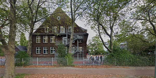 Hinter einem Zaun und hohen Bäumen steht ein Backsteingebäude mit steilem Dach. In der Nähe des Eingangs versammeln sich Menschengruppen – möglicherweise zu den Aktivitäten des Offenen Ganztags. Abgefallenes Laub bedeckt den Boden und unterstreicht die herbstliche Atmosphäre.