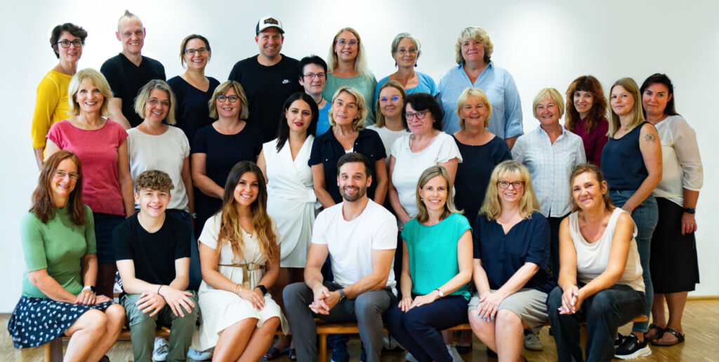 Eine Gruppe von 27 Erwachsenen unterschiedlichen Geschlechts und Alters posiert gemeinsam in einem Raum vor einer schlichten weißen Wand. Einige sitzen, andere stehen hinter ihnen und lächeln in die Kamera.