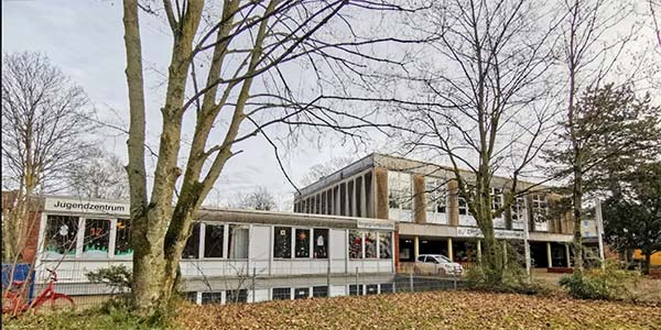 Ein modernes Gebäude mit großen Fenstern, umgeben von kahlen Bäumen und einem mit Laub bedeckten Rasen. Ein Schild mit der Aufschrift „Jugendzentrum“ weist darauf hin, dass es sich um ein Jugendzentrum mit Kinderbetreuung handelt.
