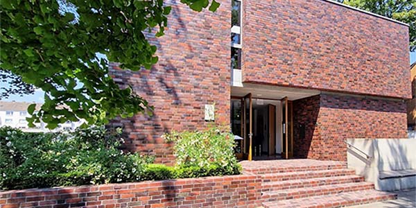 Ein modernes Gebäude mit einer Fassade aus rotbraunen Ziegeln, klaren Linien, einem zurückversetzten Eingang und einer angelegten Grünanlage davor, die teilweise von Ästen beschattet wird – ideal für Kindertageseinrichtungen oder Kindergärten.